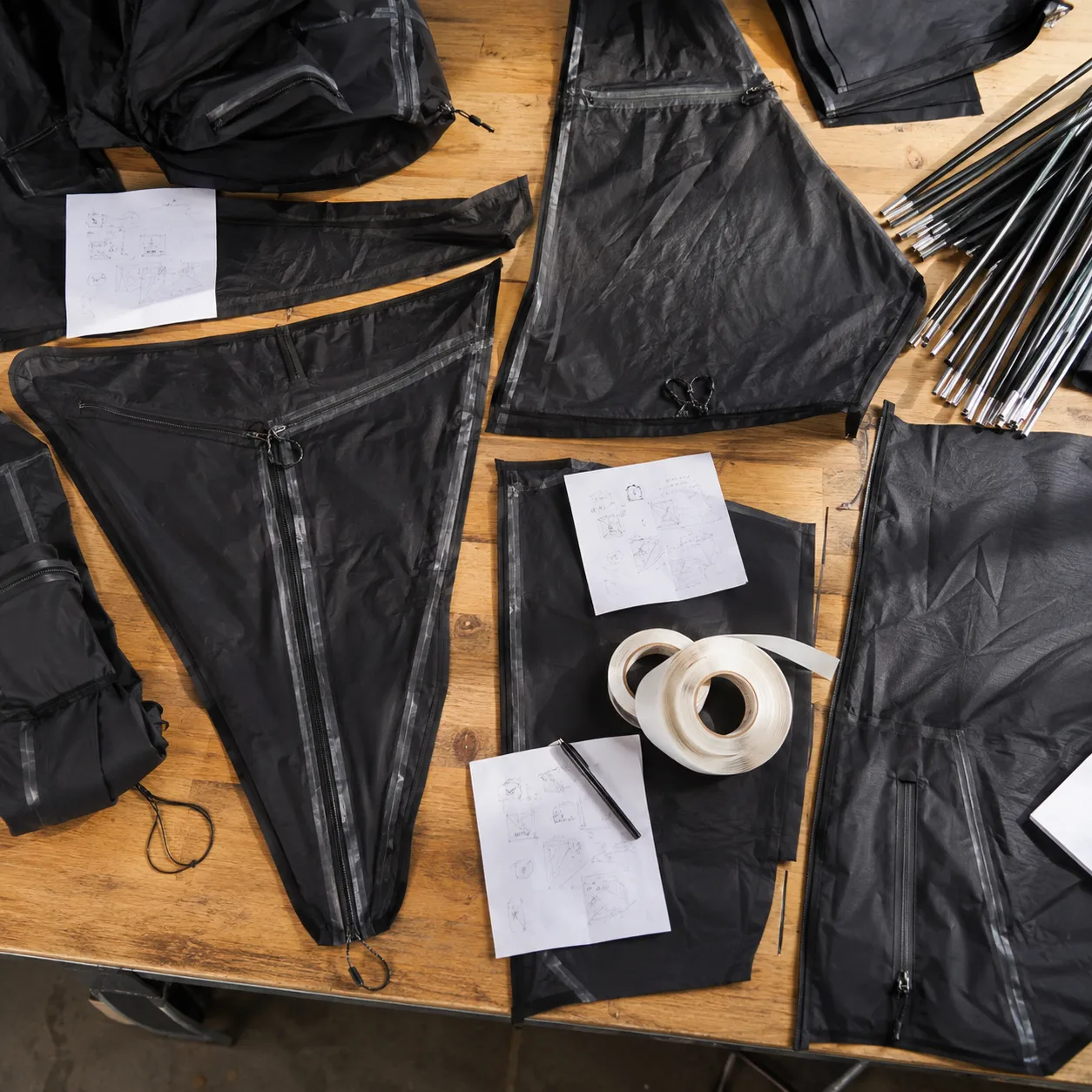 Overhead worktable with black tent pattern panels, zippers, poles, and notes under crisp angled light.