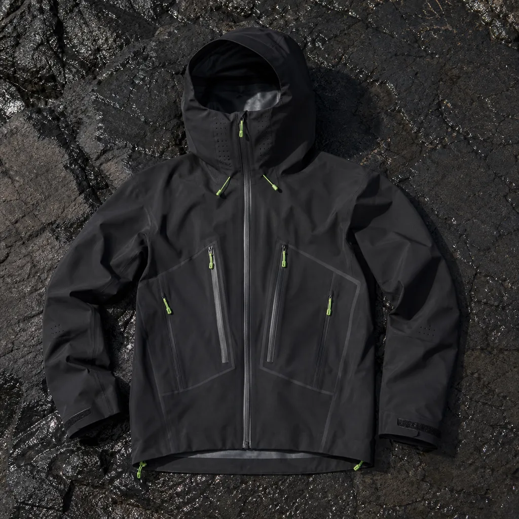 Black shell jacket laid flat on basalt rock with taped seam lines, waterproof zipper, and high hood shape visible.