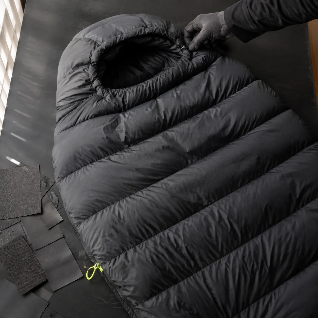 Deep charcoal mummy sleeping bag partly unrolled on a technical material table.